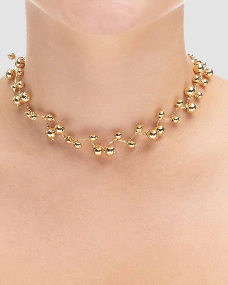 choker bolinhas dourada Papoula sendo usada por modelo, semijoia banhada a ouro 18k com design em bolinhas delicadas, 30cm + 15cm de ajuste, modelo com pele clara, blusa texturizada minimalista, estilo editorial premium, maquiagem natural elegante, fundo neutro cinza, fotografia de produto para e-commerce