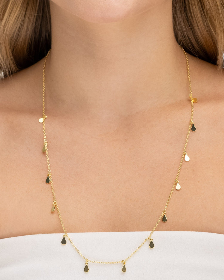 modelo usando colar dourado Papoula de 60 cm com gotas da coleção Lluvia, semijoia banhada a ouro 18k com extensor de 10 cm, fotografia destacando o caimento alongado no colo, brilho suave, movimento delicado das gotas e acabamento premium da peça