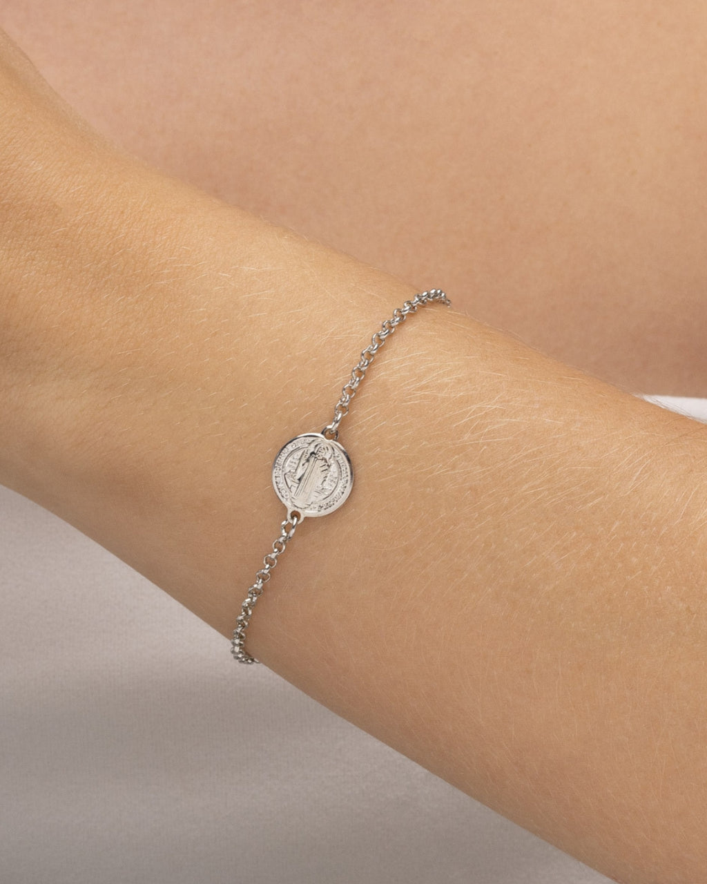Modelo usando pulseira feminina prateada com medalha São Bento no pulso — semijoia religiosa delicada Papoula