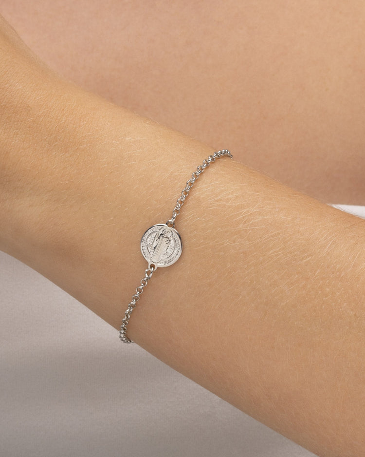 Modelo usando pulseira feminina prateada com medalha São Bento no pulso — semijoia religiosa delicada Papoula