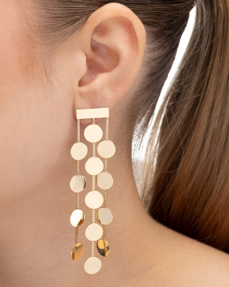 brinco dourado Papoula com design em cascata, corrente fina com discos circulares, 8 cm de comprimento e 1,5 cm de largura, semijoia com acabamento polido, fotografado na orelha