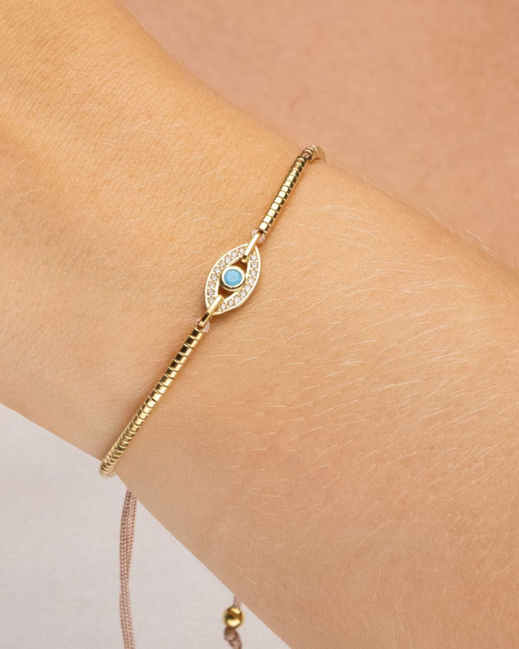 pulseira ajustável dourada com pingente de olho grego com zircônia, semijoia feminina banhada a ouro 18k usada no braço, fotografia em fundo claro com luz suave, acessório delicado de proteção e estilo moderno