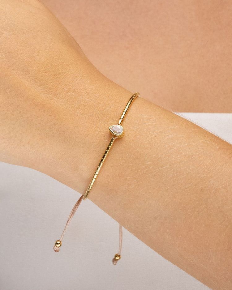 pulseira ajustável dourada Papoula com mini canutilhos e pingente de zircônia em formato de gota medindo 0,8 cm por 0,5 cm, semijoia feminina banhada a ouro 18k usada no braço, fotografia em fundo claro com luz suave destacando brilho, acabamento premium e caimento delicado da peça