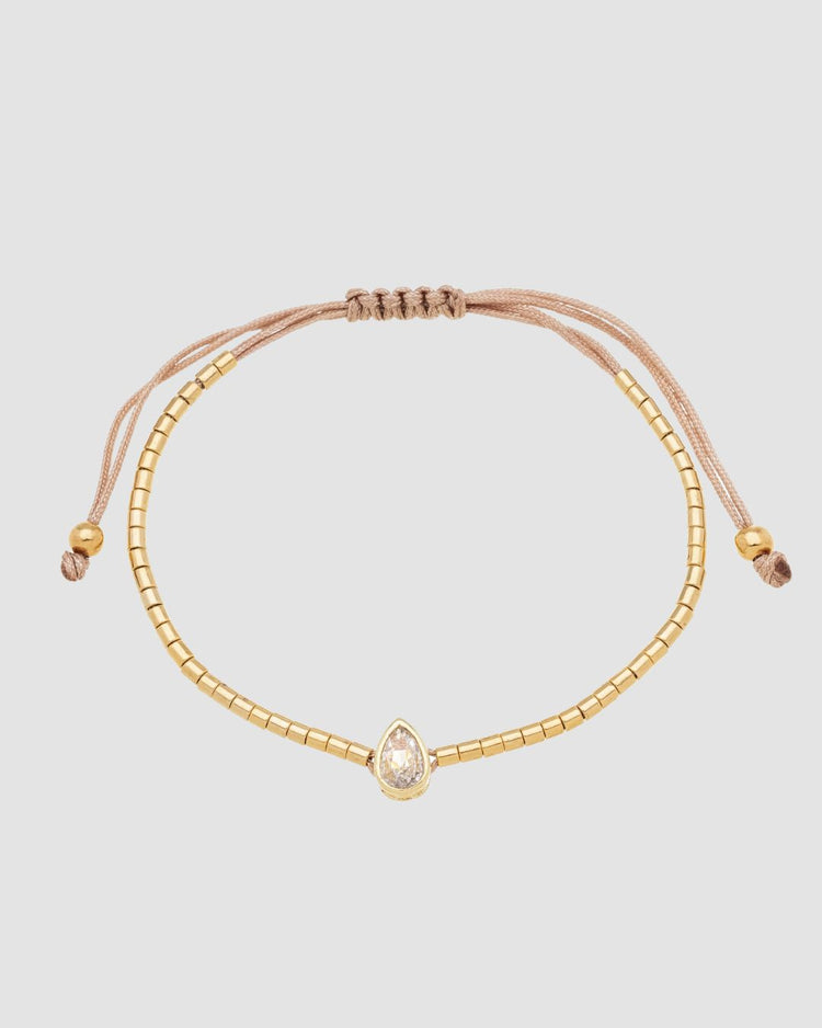 pulseira ajustável dourada Papoula com mini canutilhos e pingente de zircônia em formato de gota medindo 0,8 cm por 0,5 cm, semijoia feminina banhada a ouro 18k, acessório delicado e minimalista fotografado em fundo claro com luz suave, destaque para brilho da gota e acabamento premium da peça