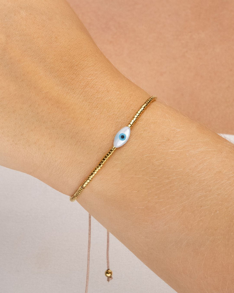 Pulseira ajustável dourada com canutilhos e pingente de olho grego oval branco e azul, banhada a ouro 18k, fotografada no pulso sobre fundo branco com sombra suave.