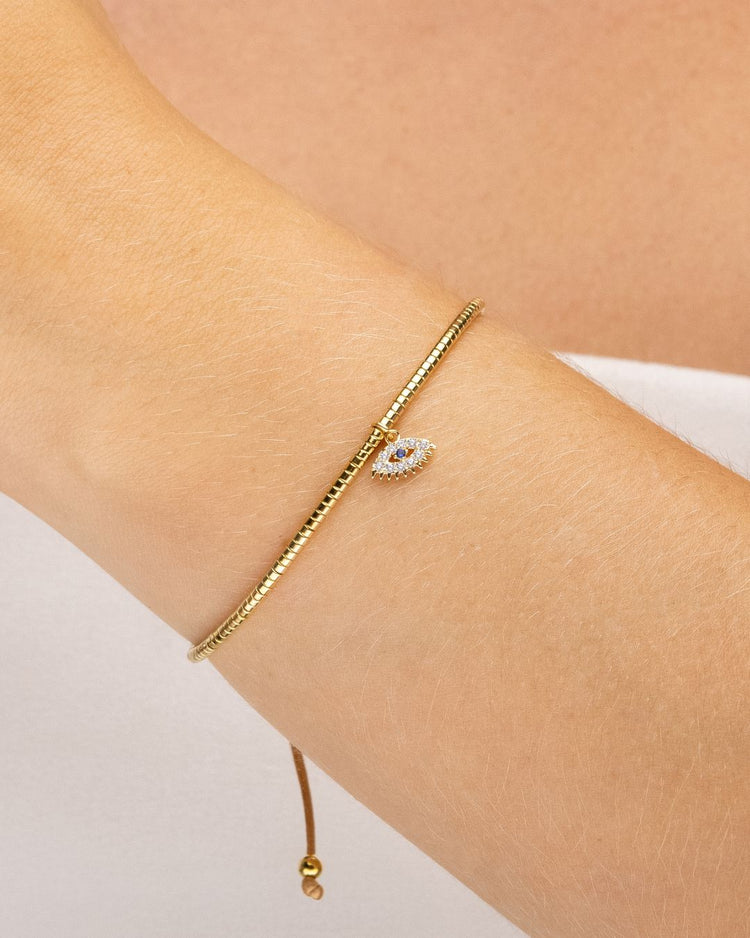 pulseira ajustável dourada com mini canutilhos e pingente de olho grego com zircônia usada no pulso, semijoia feminina banhada a ouro 18k, fotografia em fundo claro com luz suave destacando o acessório