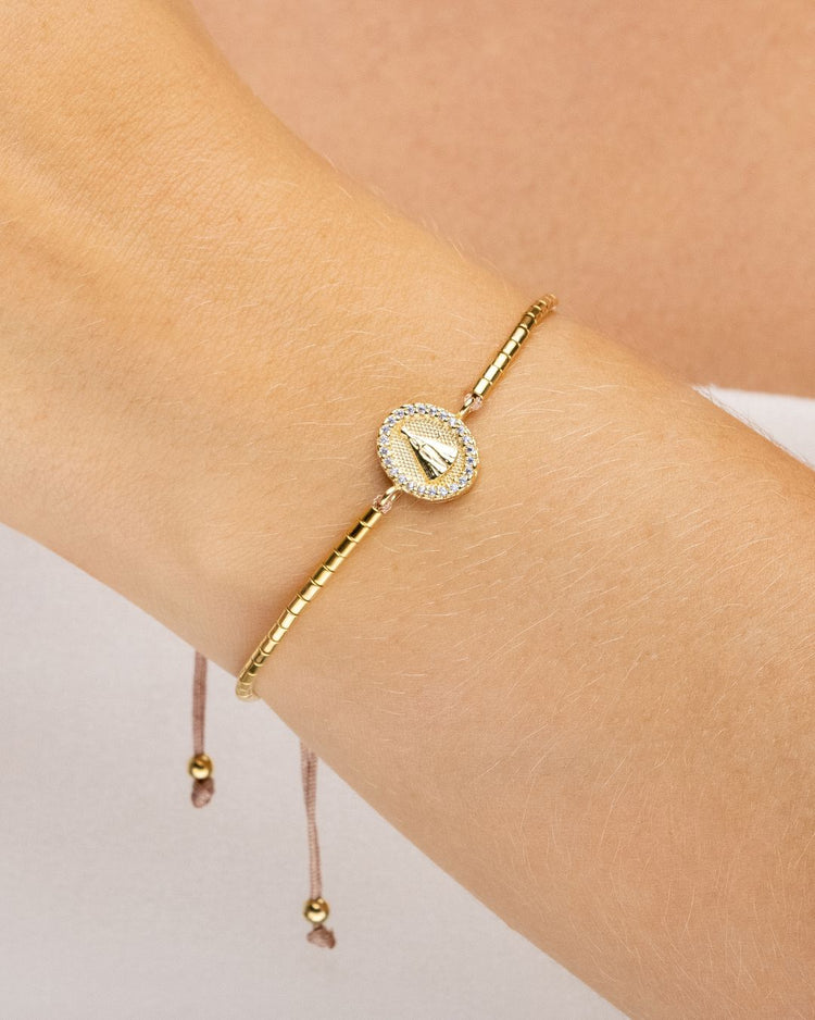 pulseira ajustável dourada com canutilhos e pingente de Nossa Senhora Aparecida com mini zircônia usada no pulso, semijoia feminina banhada a ouro 18k, fotografada em fundo claro com luz suave destacando o detalhe do pingente