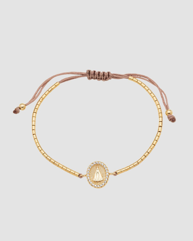pulseira ajustável dourada com canutilhos e pingente de Nossa Senhora Aparecida com mini zircônia, semijoia feminina banhada a ouro 18k, fotografia em fundo claro com luz suave destacando o detalhe religioso do pingente
